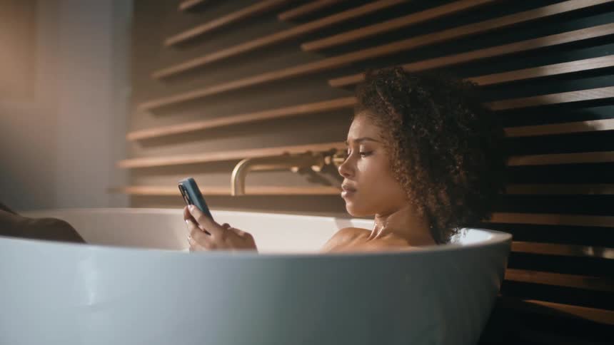 A woman is texting on her cell phone while taking a bath