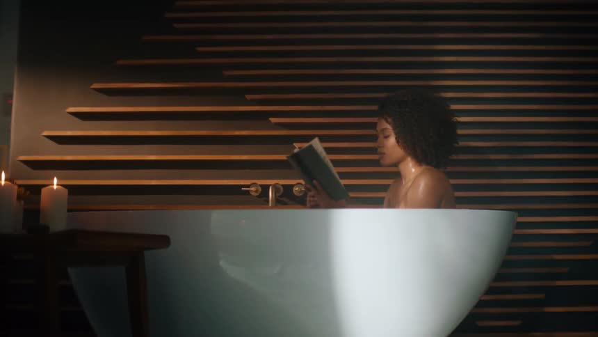 A woman in a bathtub reading a book next to candles on the table next to her