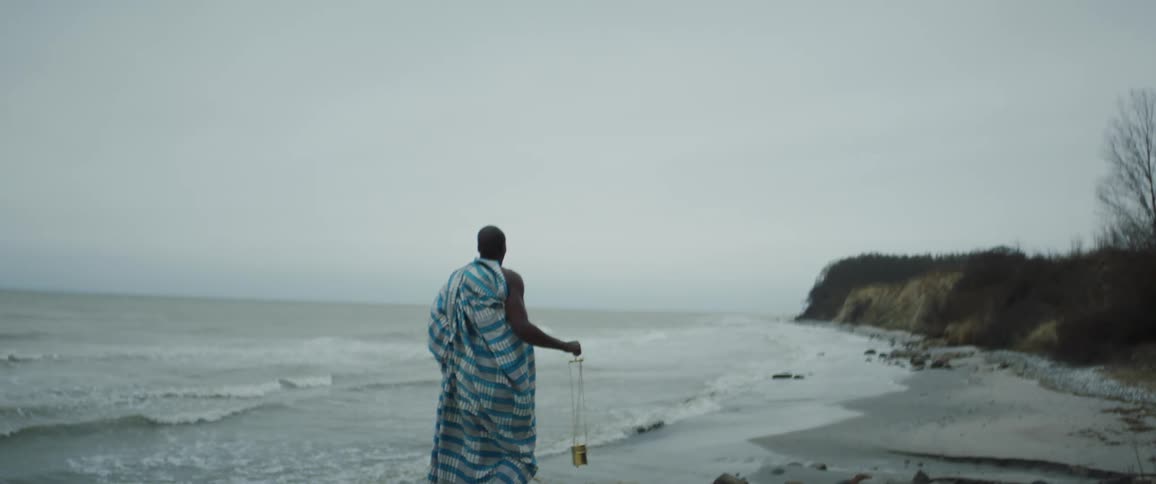 A black man wearing a striped robe is walking along the beach while carrying a gold cylinder