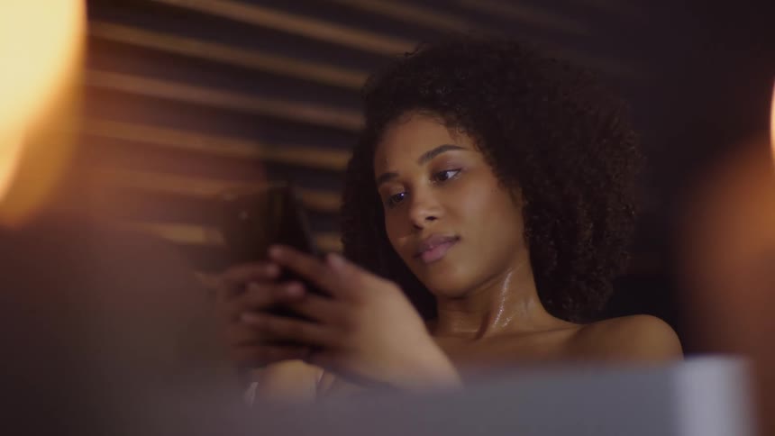 A beautiful black woman is texting on her phone while soaking in a bathtub under candlelight
