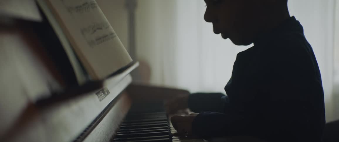 A little boy plays the piano while his father sits behind him