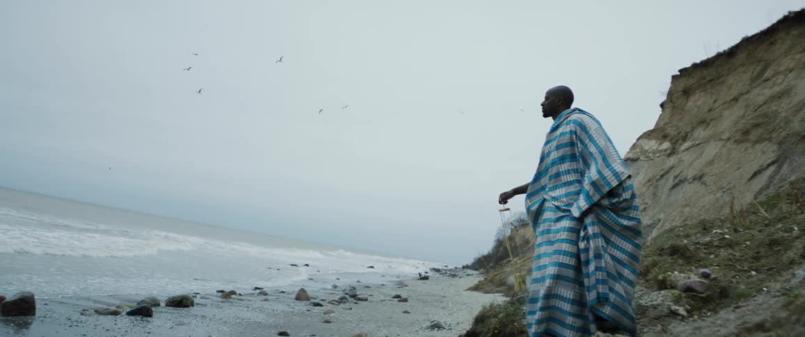 A man wearing a blue checkered cloth swinging somehting that is smoking looking out over the ocean
