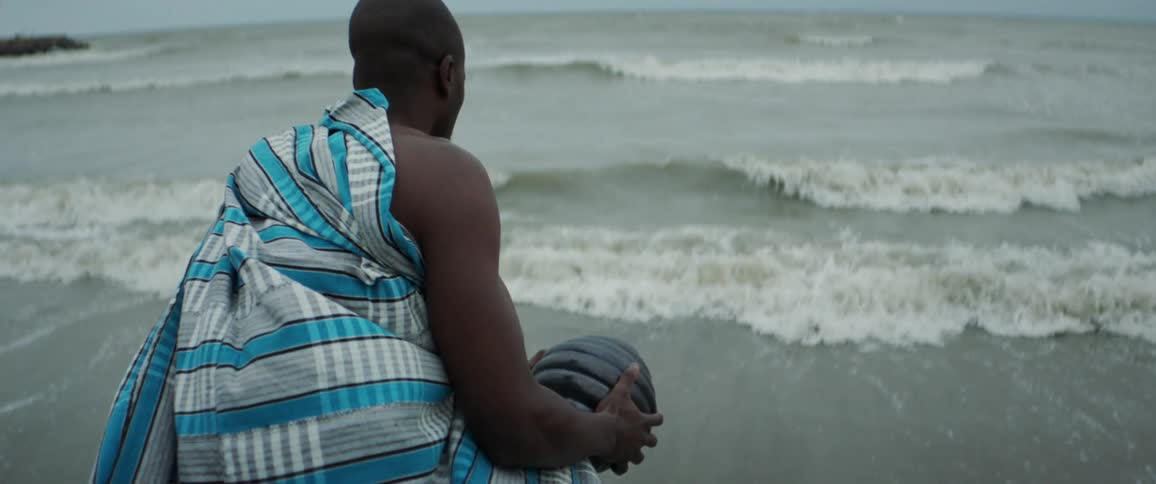 Black man wearing plaid sheet throwing ashes into the ocean from shoreline