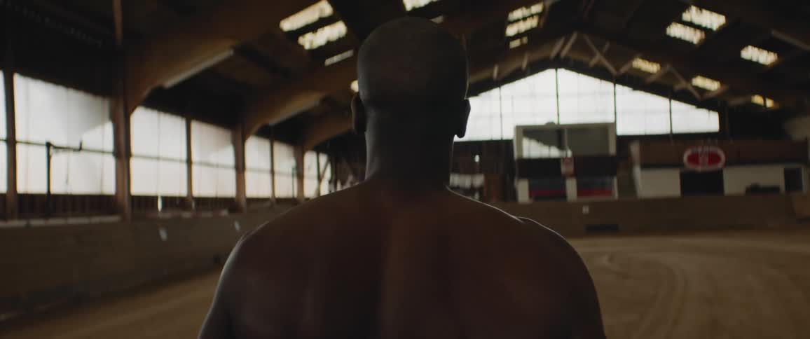 Shirtless man walks across indoor horse riding arena strewn with multiple windows