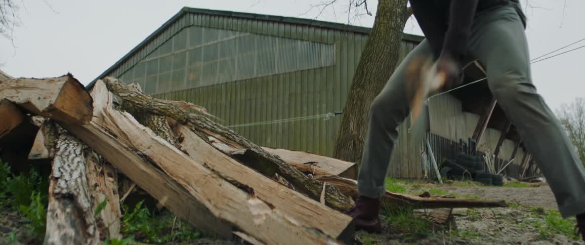 A man in a cowboy hat angrily chops at a pile of splintered wood logs with an ax