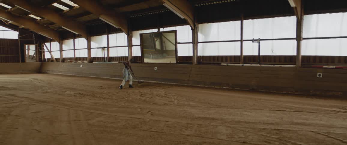 A shirtless man shovels dirt inside a large show barn in the day