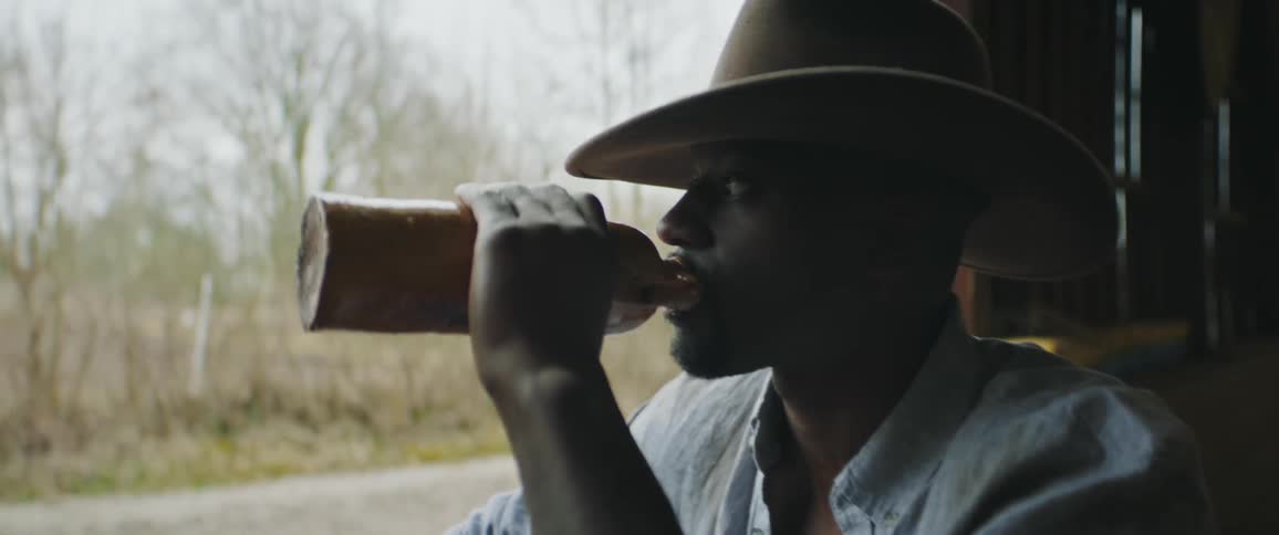 Young man wearing a cowboy hat standing on a barn drinks from a vintage ceramic bottle