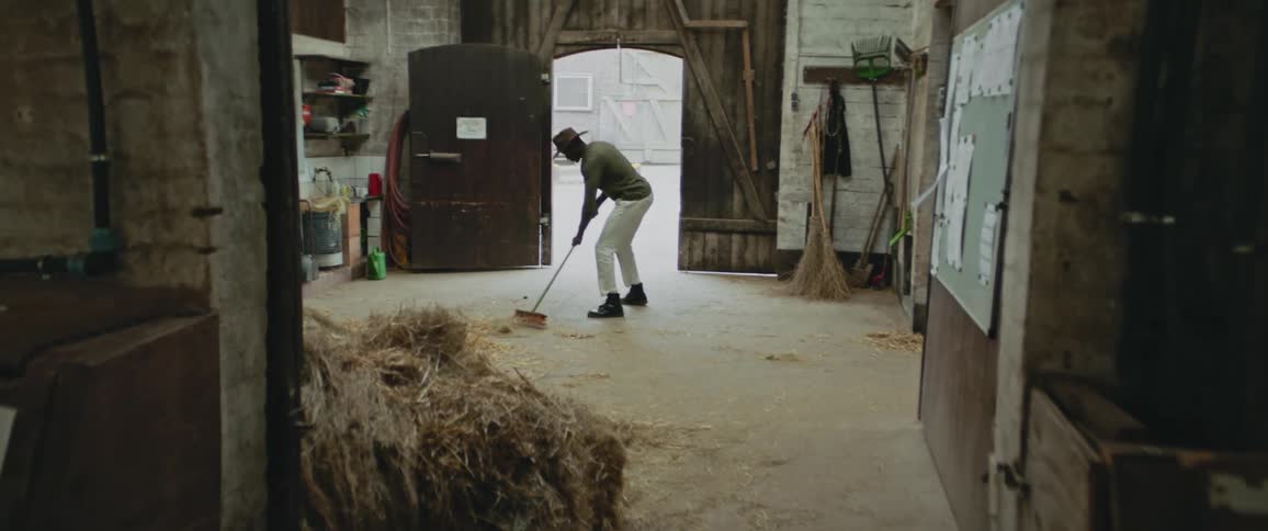 Young man wearing a cowboy hat sweeps the hay on the floor of a barn