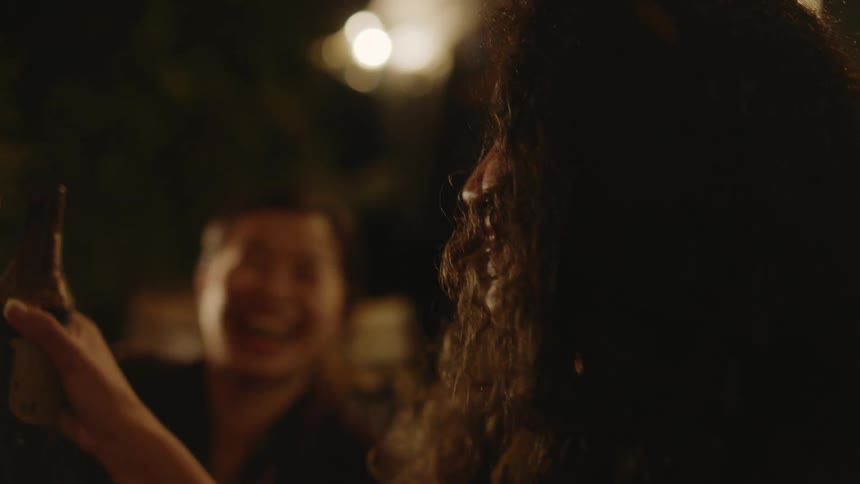 An up close of a woman laughing and smiling holding up her drink as a woman in the back does the same