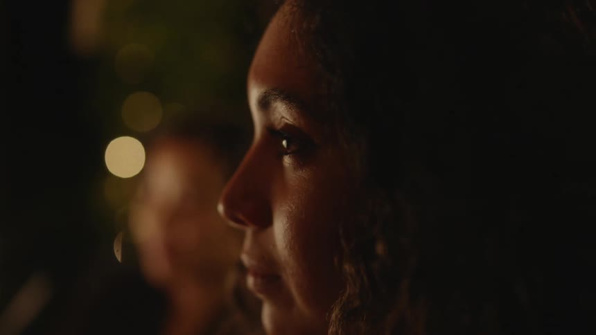 A woman is looking forward intently with a relaxed look as if she is enjoying the thing she is watching beyond the side of her face is another woman also watching in the same direction with greenery and lights in the dark outdoor area beyond her