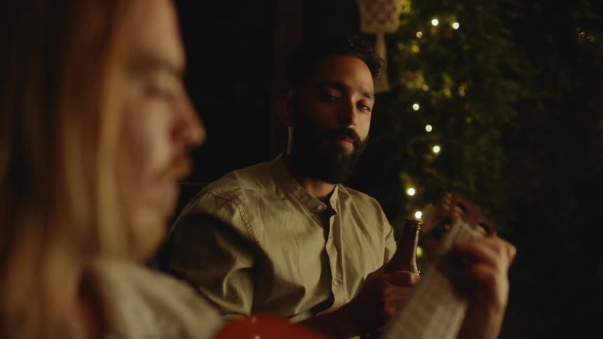 A man is sitting down playing an acoustic guitar and a man is sitting near him drinking a beer