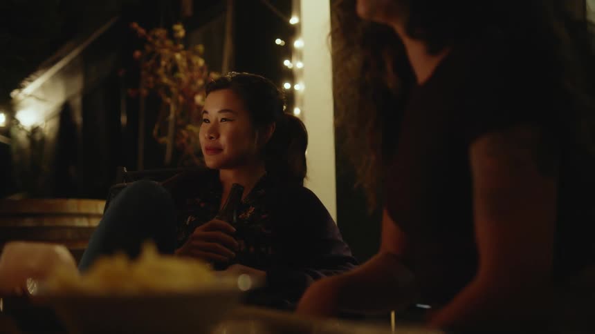A young asian woman holding a beer laughs while sitting with friends on an outdoor patio at night