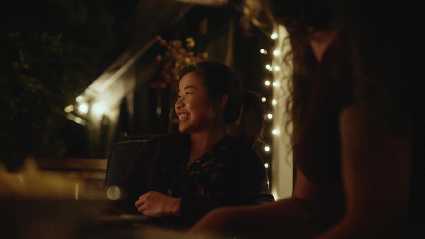 A woman is leaning her head back and laughing while conversating in an outdoor dinning area lit with garden lights at night