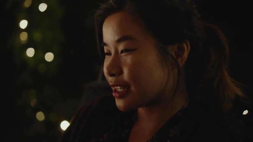 Light shines on a womans face as she speaks and lights glow in the darkness in the distance