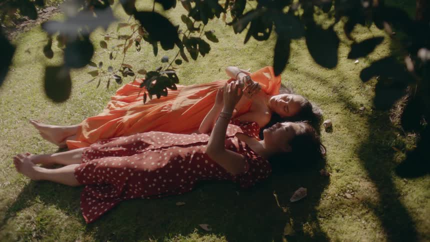 Two women laying on the grassy ground looking at objects in their hands and showing one another