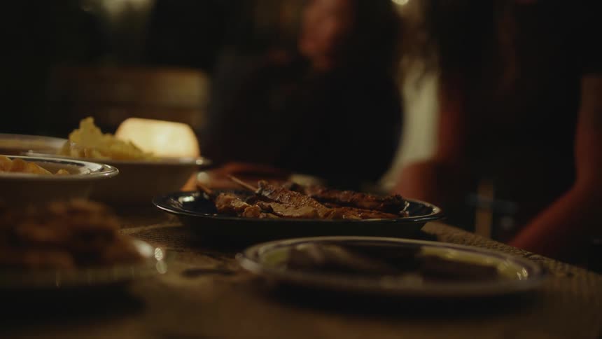 A table is set in an outdoor setting at night with bowls and plates resting on it that have food waiting to be served while people are talking and sitting down in background