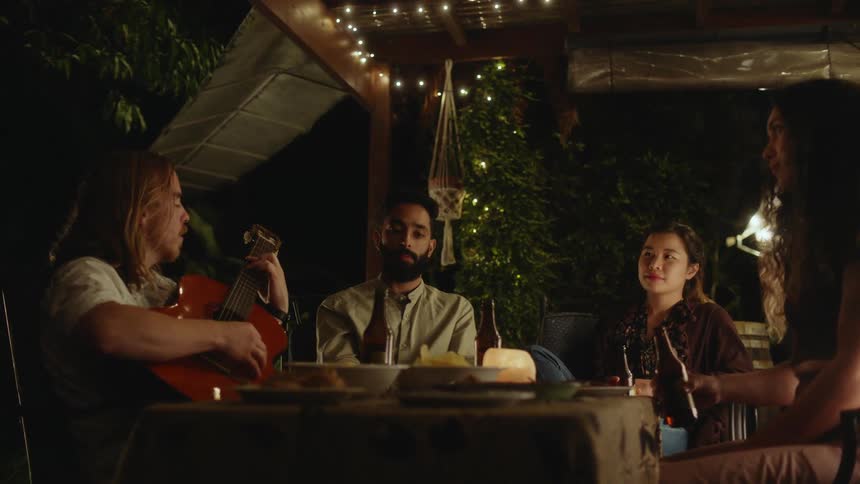 A small group of people are sitting around a table drinking beer as a man plays an acoustic guitar