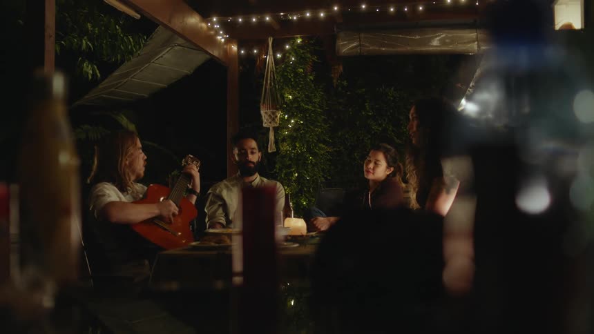 A group of people are listening to a man play the guitar while sitting around a table at night