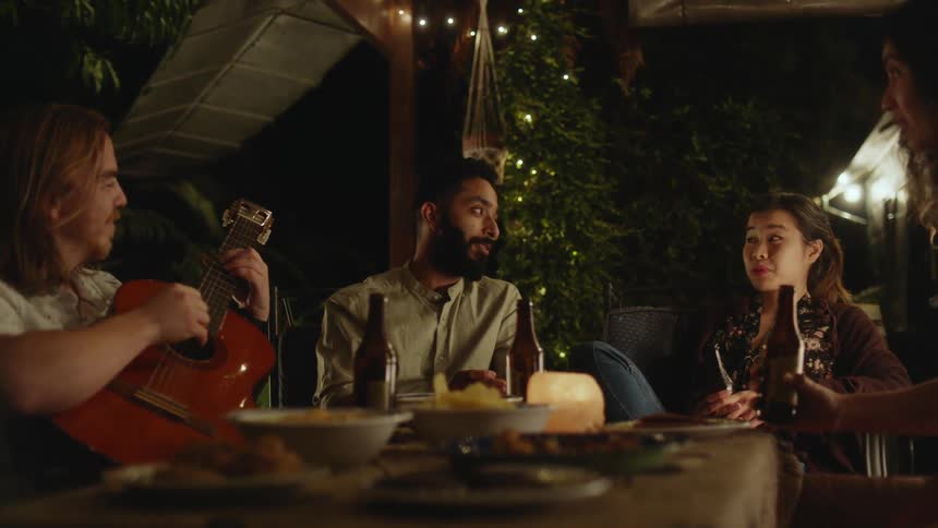 A small group of people sit around a table talking as one person plays a guitar