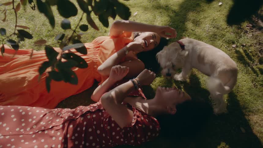 Two women laying on the ground laughing and smiling as a dog comes up to their faces