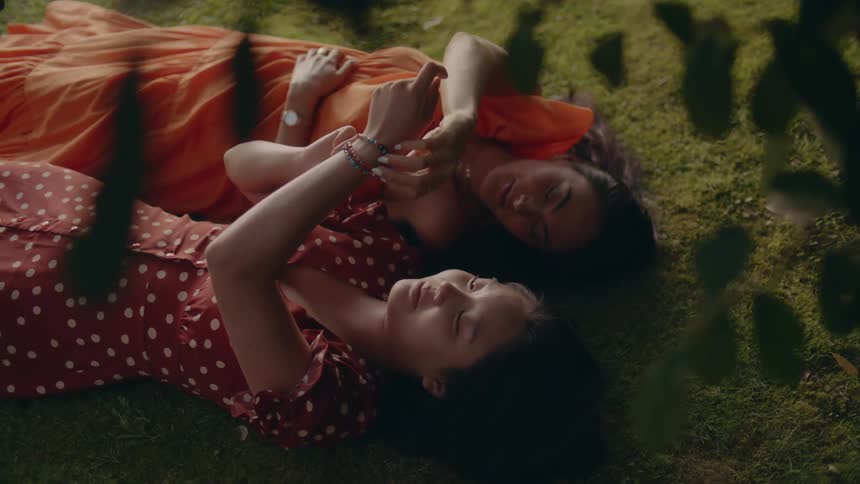 Two young girls wearing bright colored spring dresses lying on the mosscovered ground exchanging bracelets and smiling