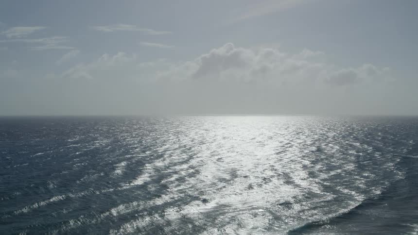 Dramatic descent over rolling ocean waves as sunshine shimmers and reflects off of the surface