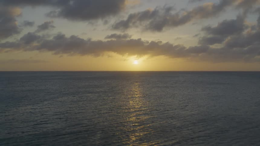 Pullback descent over calm ocean water as the low hanging sun reflects warm light from shimmering ripples and gentle waves under a painted sky