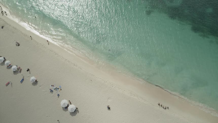 Birds eye view of a coral sand beach lapped by tranquil clear water and sunbathers with white umbrellas