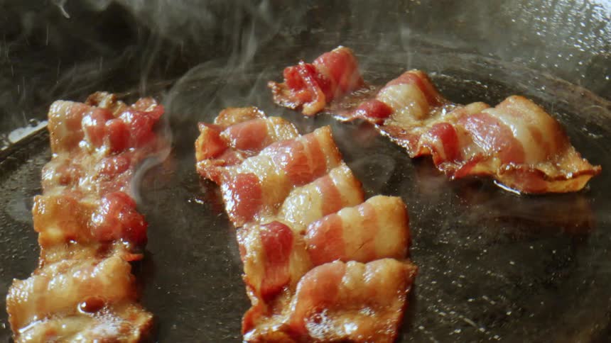 A large frying pan cooks sizzling bacon with lots of smoke emitting from it