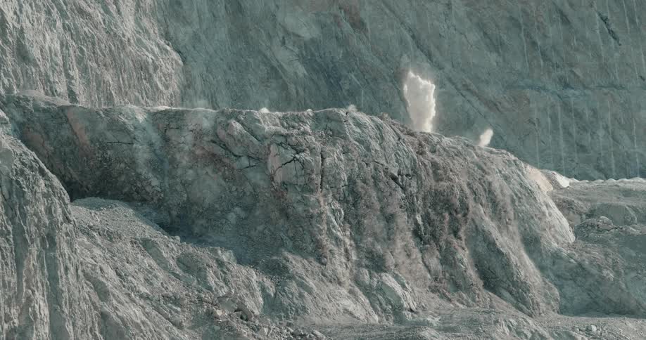 The top of a volcano covered in ash expands out as a plume of smoke rises and a black dust begins to roll down down the side of the hill