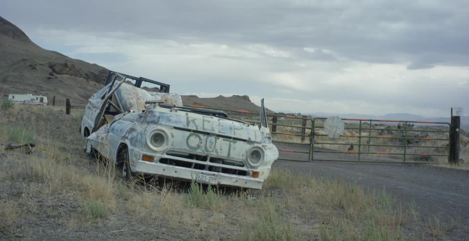 An old wrecked car sits on the side of a road near a gate in the desert with mountains in the background