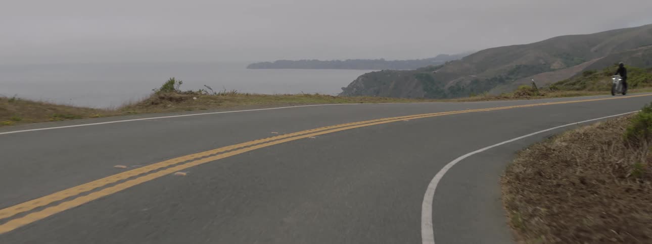 Motorcyclist drives motorbike around curving road near coast on misty day