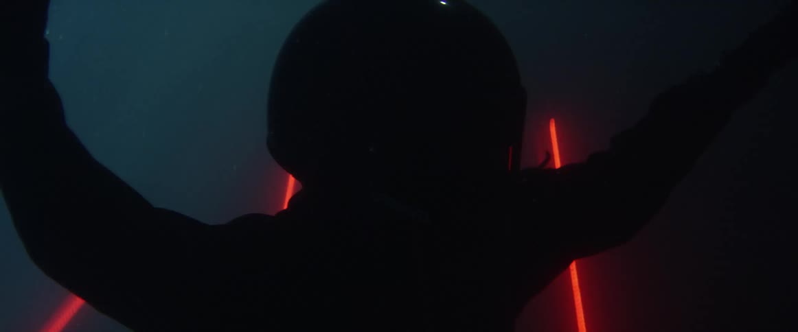 A person wearing a black suit and helmet floating in water with red lasers behind them