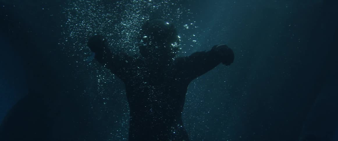 A person wearing all black and a black helmet floating under water as bubbles surround them
