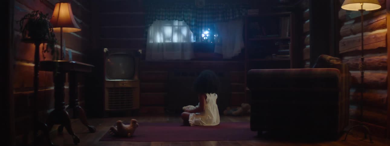 Little girl in nightgown sitting on the floor of living room of cabin in the woods with teddy bear car headlights approaching in the window