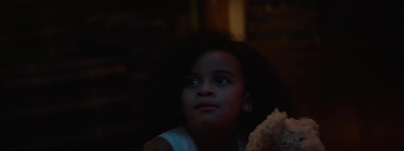 A little girl is sitting down holding a teddy bear and watching a tv that is off camera