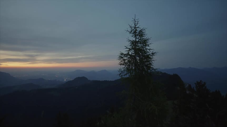 A tree stands on top of a mountain as the sun is setting in a hazy sky and there are mountains in the distance