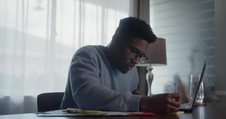 A man is sitting at a desk with a laptop computer as he writes on a notepad