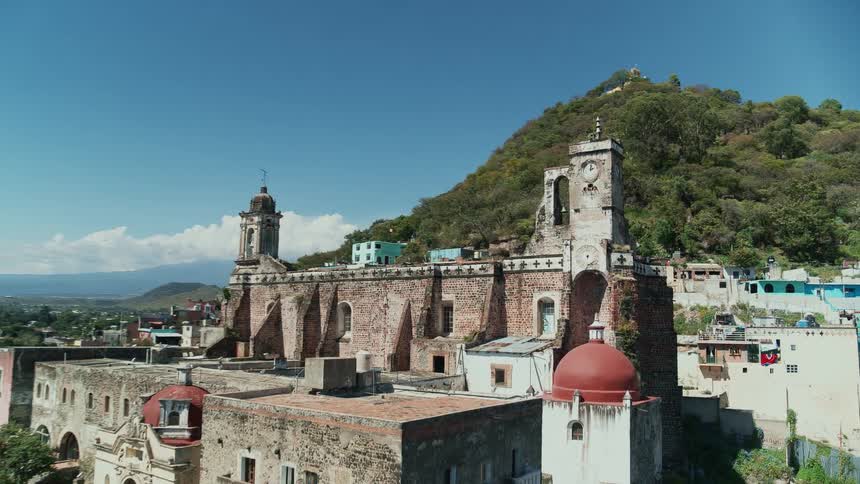 An old brick building with a wall and a clocktower stand near buildings along the foot of a mountain overlooking a city
