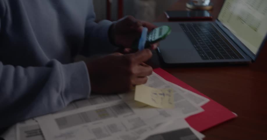 A young man sits at a desk and uses a calculator while writing on a sheet of paper