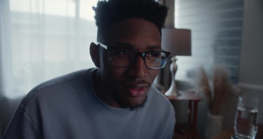 Man with glasses and blue sweater sits at desk thinking and writing down in notebook in room