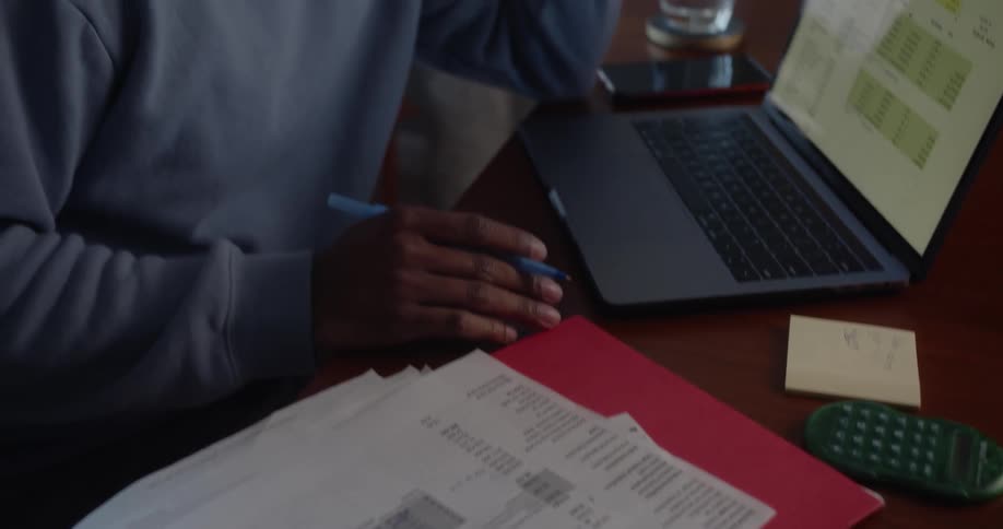 Man with blue sweater sits at table working on documents and laptop