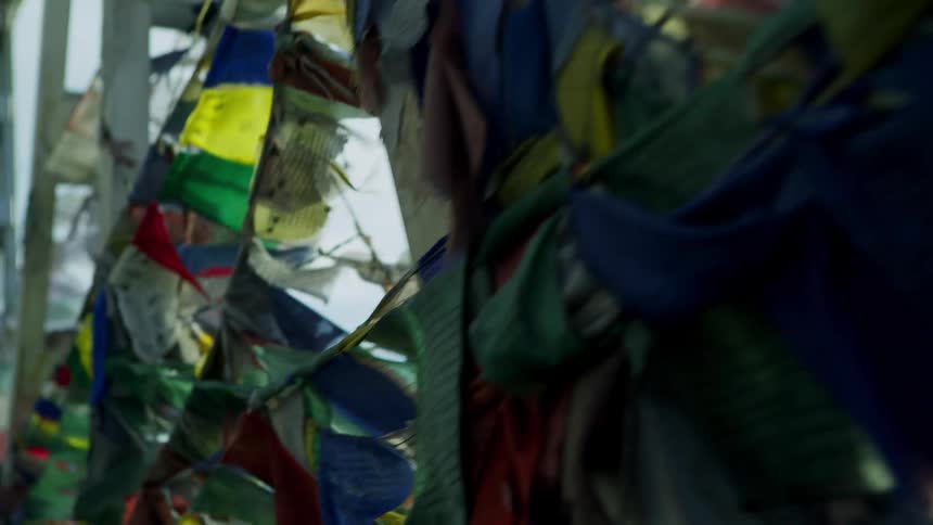 Many colored flags strung across a bridge waver in the wind and there are mountains in a foggy sky in the distance