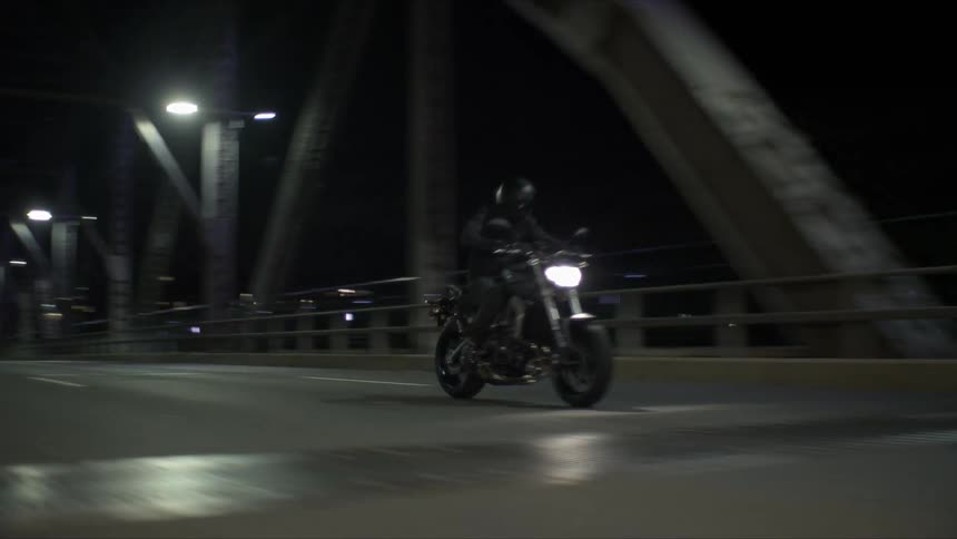 A motorcycle with the headlight on crosses a bridge as traffic moves in the distance