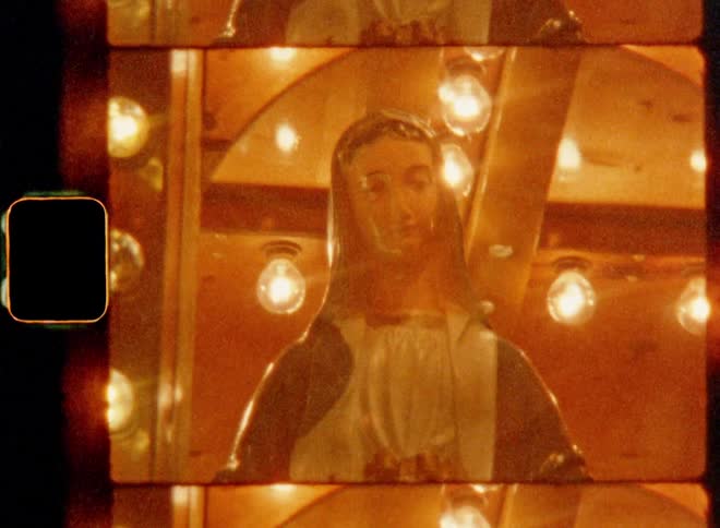 Statue of person in pastoral attire resembling the Blessed Virgin Mary spins slowly on a Christmas pyramid or merry-go-round illuminated by the warm shine of many electric light bulbs on vintage 8mm film footage with smooth sidewise movement