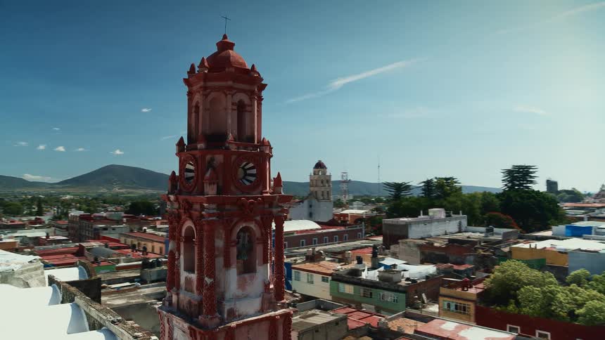 Bell tower stands over colorful buildings near road in town bordering mountains