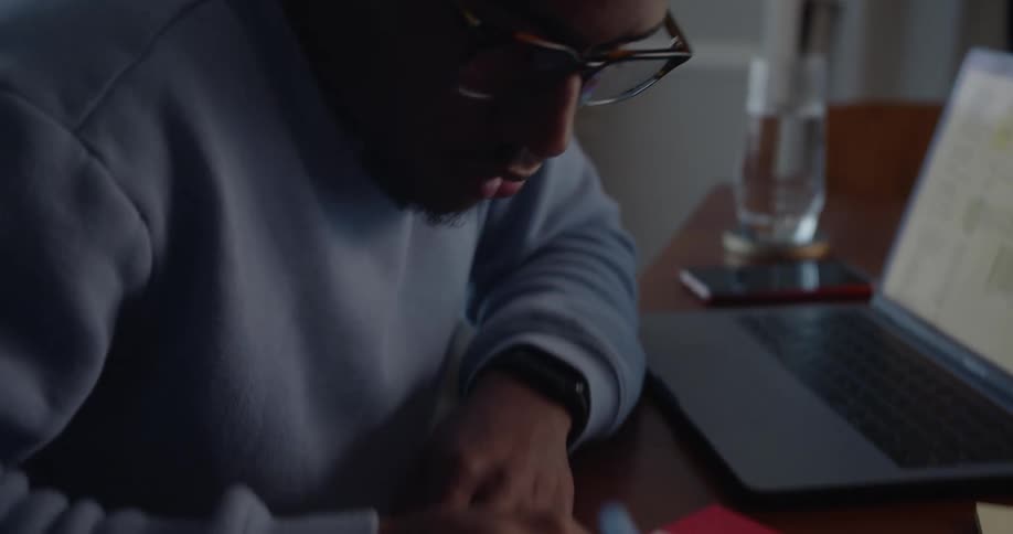 A male sits at a desk while concentrating as he writes on a document with numerical tables on it