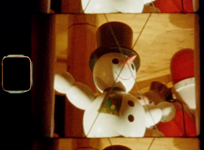 A snowman sculpture with a carrot nose and a top hat and a smile on his happy face next to a Father Christmas figure turns on a holiday carousel display in the dark in front of black night sky illuminated by many electric light bulbs
