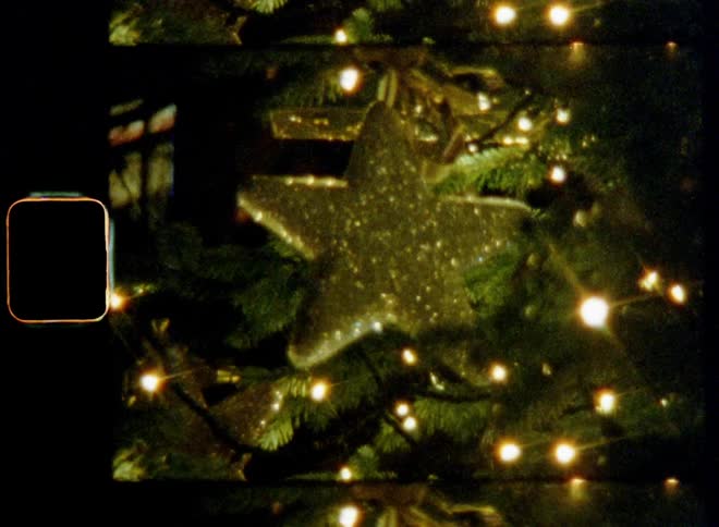 Handheld dynamic 8mm film footage of glittering and flashing golden star ornament with reflective surface covered in glitter that sparkles brightly as it hangs on Christmas tree with pine needles lights and candles that shine bright with lens flare