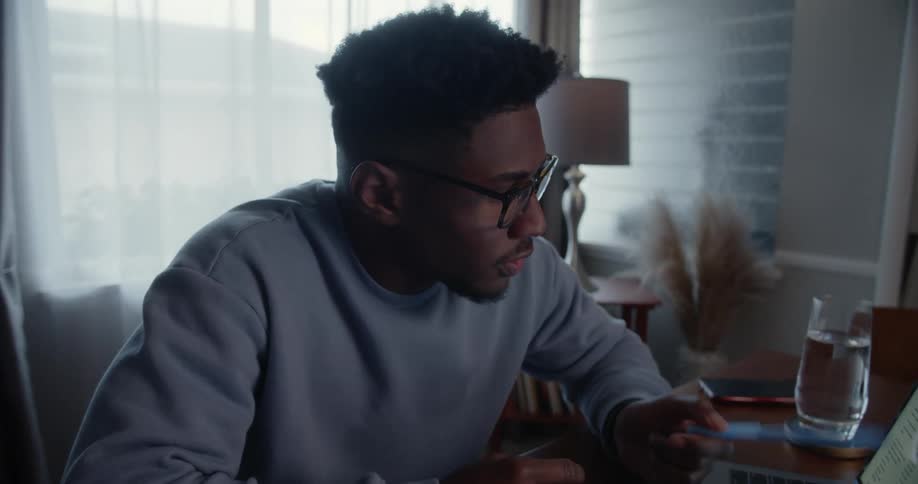 Man with glasses wearing blue sweater works on laptop in room